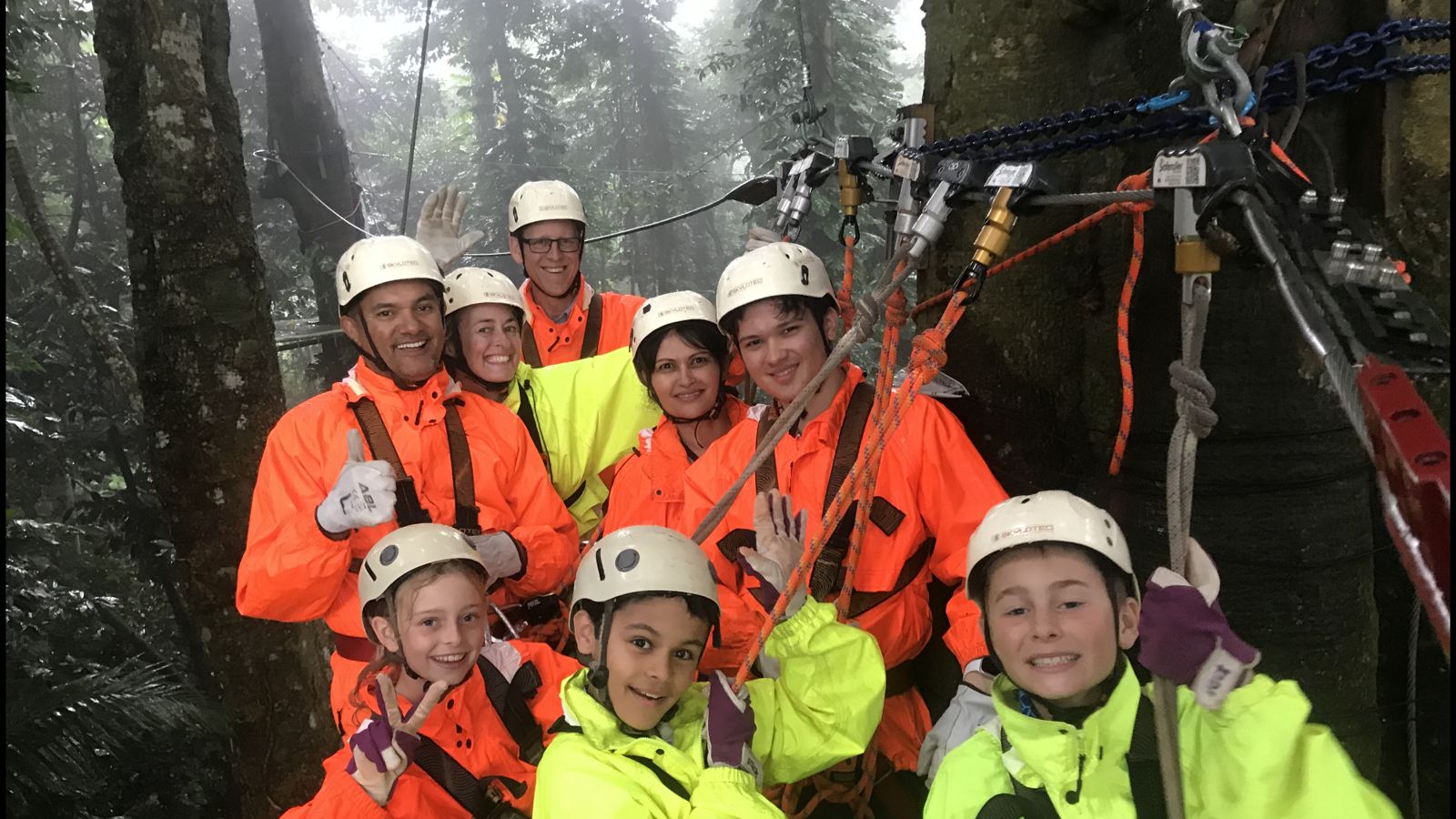 Un groupe souriant en vestes de pluie et casques sur une plateforme de tyrolienne dans la forêt tropicale brumeuse